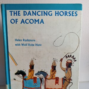 May include: A book cover with a blue background and the title "The Dancing Horses of Acoma" in white text. The cover features an illustration of three Native American people riding horses, with a rainbow in the background.