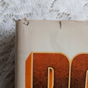 May include: Close-up of a book corner with a torn edge. The book has a cream-colored cover with large, orange and brown lettering. The background is a white lace fabric.