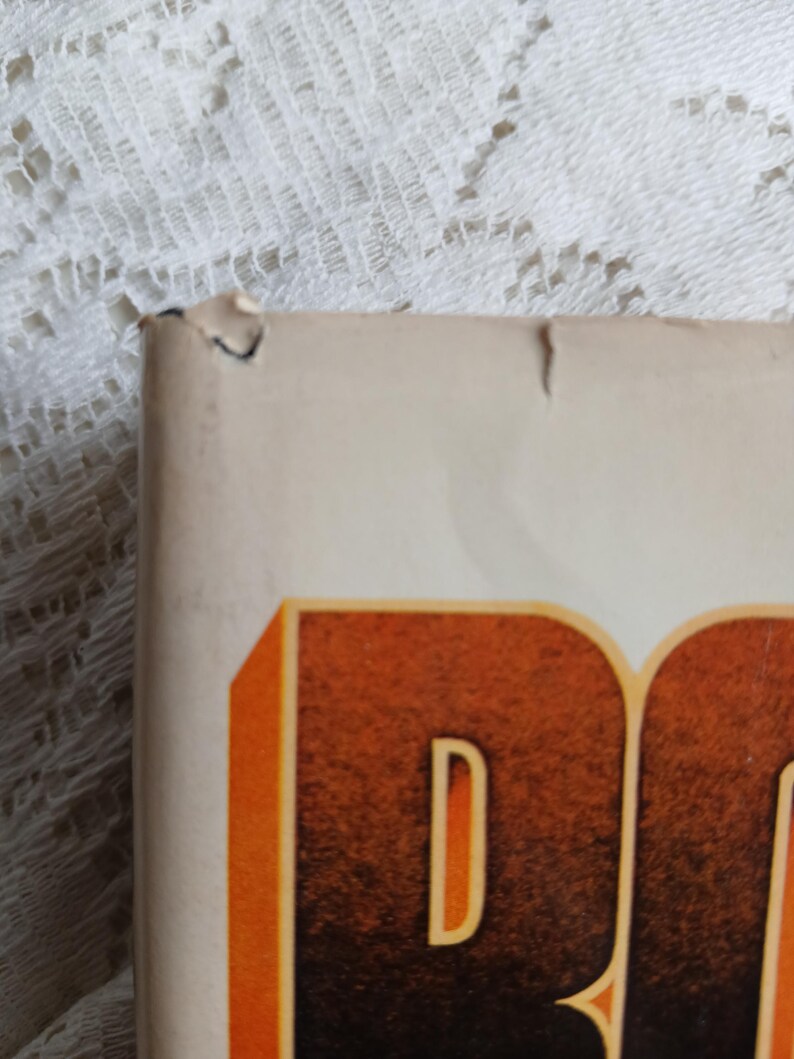 May include: Close-up of a book corner with a torn edge. The book has a cream-colored cover with large, orange and brown lettering. The background is a white lace fabric.