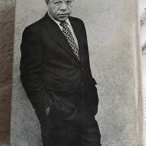 May include: A black and white photograph of a man in a dark suit, white shirt, and patterned tie. He wears glasses and stands with his hands in his pockets. The background is a mottled gray.