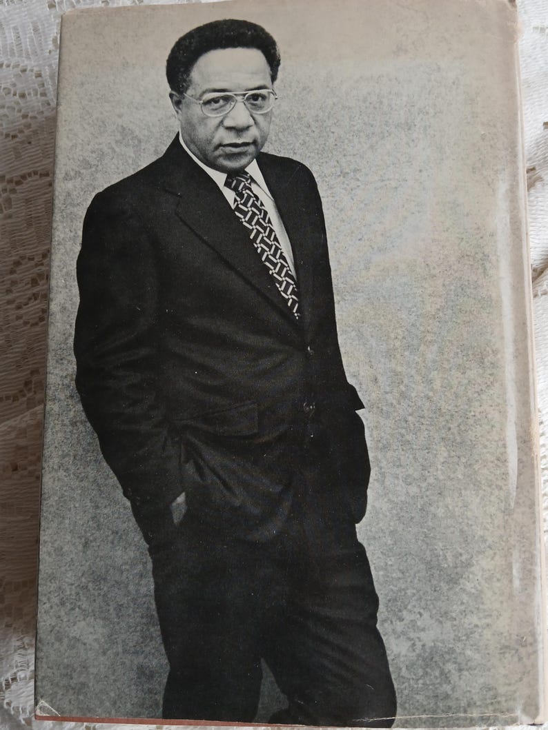 May include: A black and white photograph of a man in a dark suit, white shirt, and patterned tie. He wears glasses and stands with his hands in his pockets. The background is a mottled gray.