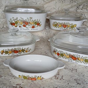 May include: A set of five white ceramic casserole dishes with glass lids. Each dish features a colorful floral and vegetable design with text. The dishes vary in size and shape, suitable for baking and serving.