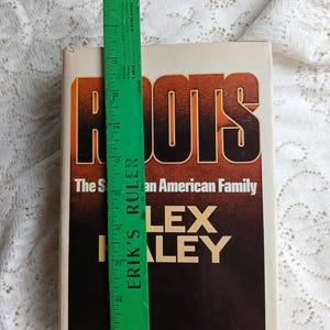 May include: A hardcover book titled "Roots" by Alex Haley. The cover features the title in large orange and black letters. A green ruler, approximately 12 inches long, is placed next to the book.