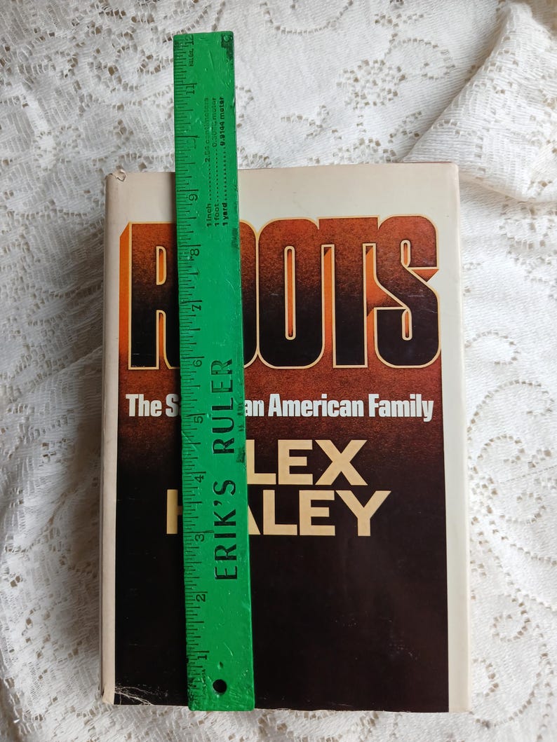 May include: A hardcover book titled "Roots" by Alex Haley. The cover features the title in large orange and black letters. A green ruler, approximately 12 inches long, is placed next to the book.