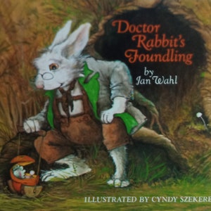 May include: A white rabbit wearing a green jacket and brown pants holds a basket with a small mouse inside. The book cover reads "Doctor Rabbit's Foundling" by Jan Wahl, illustrated by Cindy Szekerez.