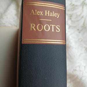May include: A hardcover book titled "Roots" by Alex Haley. The book has a dark blue cover with a brown label featuring gold lettering. The title and author's name are prominently displayed.