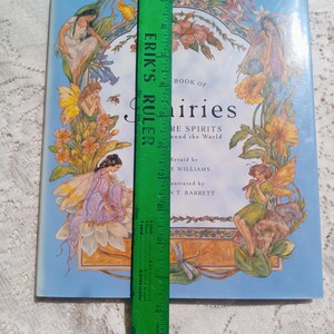 Puede incluir: Un libro de tapa dura titulado "Book of Fairies" con ilustraciones de hadas y flores. El libro es de color azul claro con una regla verde que mide aproximadamente 23 cm de largo. La portada presenta hadas y elementos florales.