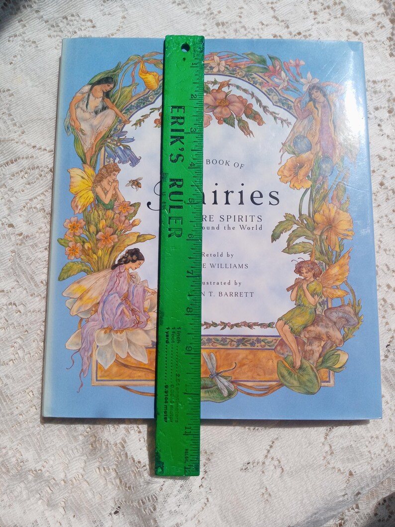 Puede incluir: Un libro de tapa dura titulado "Book of Fairies" con ilustraciones de hadas y flores. El libro es de color azul claro con una regla verde que mide aproximadamente 23 cm de largo. La portada presenta hadas y elementos florales.