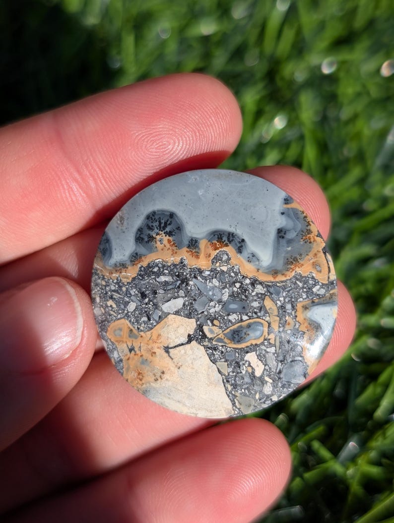 Puede incluir: Una piedra pulida y redonda con un patr&oacute;n &uacute;nico. La secci&oacute;n superior es de color gris claro con inclusiones oscuras en forma de &aacute;rbol. El centro presenta una banda de color beige y marr&oacute;n, mientras que la parte inferior es una mezcla de gris, beige y blanco, con un aspecto agrietado.