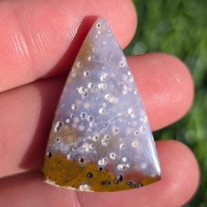 Puede incluir: Un cabujón de ágata de forma triangular, de color azul claro con un patrón blanco y marrón. La piedra tiene un aspecto moteado con pequeños puntos blancos en todo.
