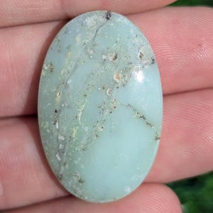 Puede incluir: Gema ovalada de color verde pálido con superficie pulida. La piedra presenta vetas sutiles e inclusiones pequeñas de color marrón oscuro, creando una apariencia natural y texturizada. Probablemente utilizada para joyería o decoración.