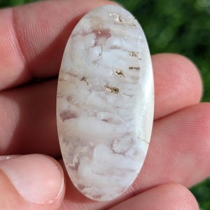Könnte beinhalten: Weißer Achat-Cabochon mit einem wirbelnden Muster und braunen Einschlüssen. Der Cabochon ist oval geformt und hat eine glatte, polierte Oberfläche.