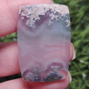 May include: A rectangular gemstone with a polished surface. The stone features a blend of colors, including pink, white, and gray, with a pattern resembling clouds and small, dark, tree-like formations. The top edge has a textured, light-colored pattern.