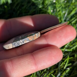 Puede incluir: Una piedra pulida de forma ovalada con una mezcla de colores grises, verdes y beige. Una moneda de plata est&aacute; al lado de la piedra. Los objetos se sostienen en una mano, con un fondo verde borroso.