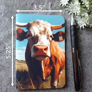 May include: A 3.5" x 5.25" notebook featuring a detailed illustration of a cow with brown and white fur, set against a blue sky and field background. A black pen rests beside the notebook. The notebook's design showcases a rural, farm theme.