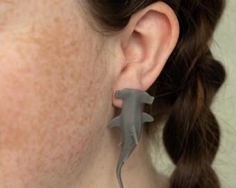 May include: A gray hammerhead shark earring. The shark is made of a smooth, gray material and has a realistic design. The earring is worn in the ear of a person with long, brown hair.