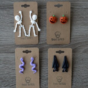 May include: Four pairs of Halloween-themed earrings on brown cardboard tags. The earrings include white skeletons, orange jack-o'-lanterns, purple snakes, and black bats. Each tag has the text "THAT BITES" printed on it.