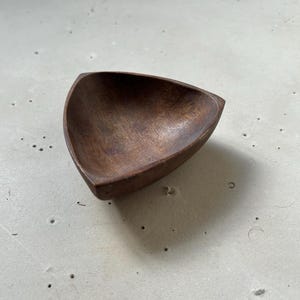 May include: A small, triangular wooden bowl with a rich, dark brown color. The bowl has a smooth, polished interior and a slightly rougher exterior. The design is simple and elegant, suitable for serving small portions of food or as a decorative item.