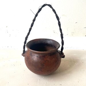Può includere: Un piccolo vaso rotondo di argilla marrone con un manico di corda scura. Il vaso ha un'ampia apertura e due piccole maniglie sui lati. Il vaso è probabilmente fatto a mano e ha un aspetto rustico.