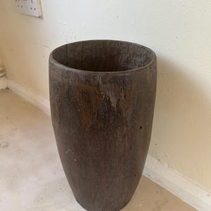 Può includere: Un alto recipiente cilindrico intagliato in legno marrone scuro con una superficie ruvida e usurata. Il contenitore presenta un'ampia apertura superiore e una base solida e arrotondata, che mostra venature naturali del legno e segni di usura.
