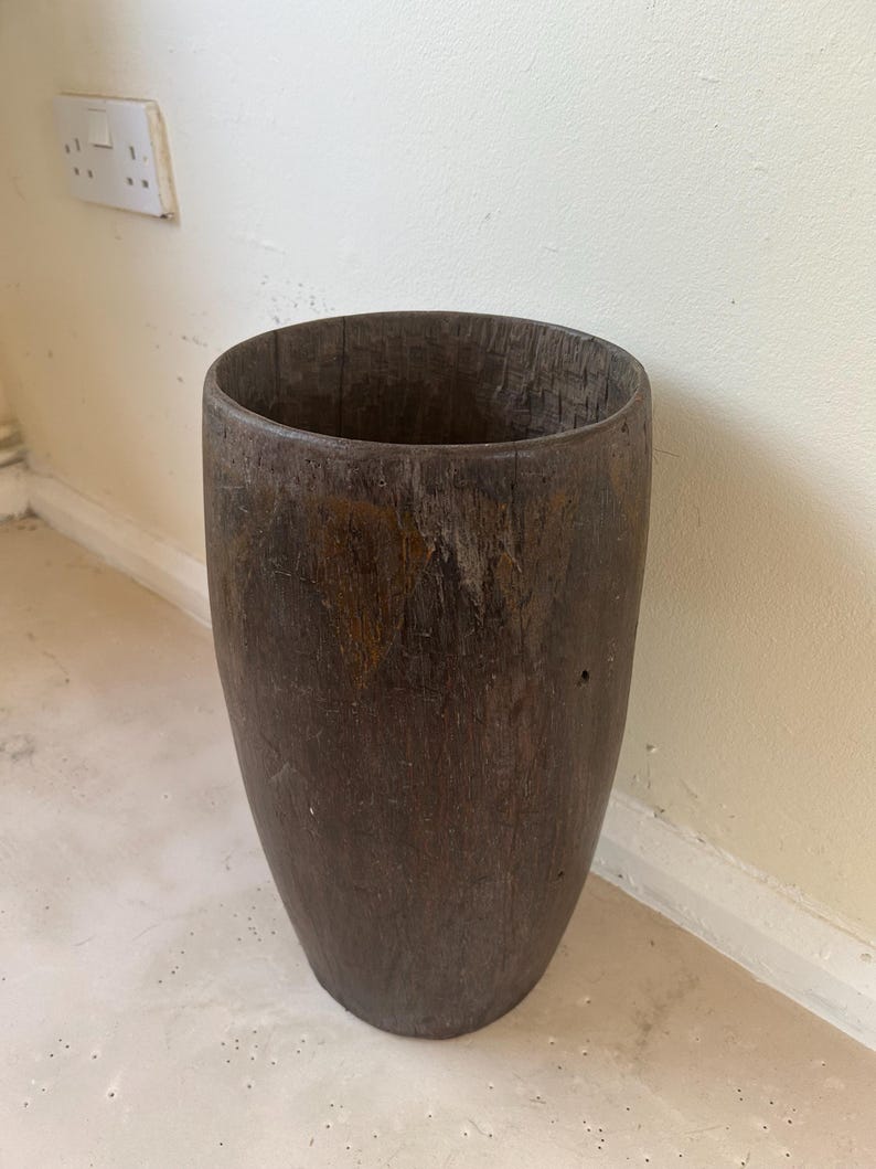 K&ouml;nnte beinhalten: Ein hohes, zylindrisches Gef&auml;&szlig; aus dunkelbraunem Holz mit einer verwitterten, strukturierten Oberfl&auml;che. Der Beh&auml;lter hat eine weite, offene Oberseite und einen soliden, abgerundeten Boden, der nat&uuml;rliche Holzmaserungen sowie sichtbare Altersspuren zeigt.
