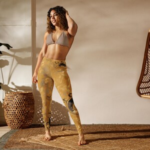 Puede incluir: Una mujer con un sujetador deportivo marrón claro y leggings amarillos con un patrón abstracto negro. Los leggings tienen cintura alta y se ajustan a las piernas. La mujer está de pie sobre una estera tejida marrón.