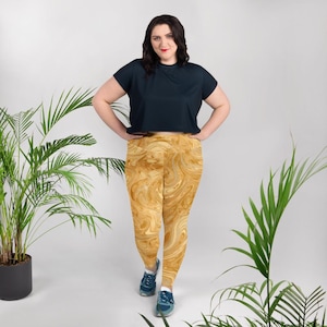May include: A woman wearing a navy blue crop top and gold-colored leggings with a swirling pattern. The leggings are high-waisted and fitted. The woman is standing in front of a white background with green plants on either side.