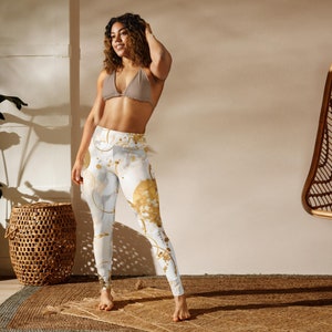 May include: A woman wearing white leggings with a gold abstract print and a brown bikini top. She is standing on a woven rug in a room with a wicker chair.