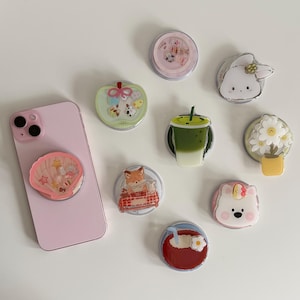 May include: A pink smartphone with a seashell phone grip, alongside several decorative phone grips. The grips showcase designs including a bunny, flowers, a fox, a bear, a matcha drink, and other charming illustrations.