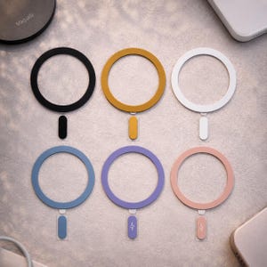 May include: Six coloured MagSafe charging rings in black, mustard, white, blue, purple, and pink. Each ring has a matching rectangular base. The word "Magsafe" is visible on a black circular charging pad.