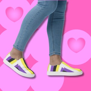 Puede incluir: Un par de zapatos de lona sin cordones con un patrón de rayas amarillas, negras y moradas. Los zapatos los lleva una persona que lleva pantalones vaqueros azules.