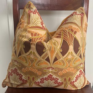 Puede incluir: Coj&iacute;n decorativo con un dise&ntilde;o floral Art Nouveau en tonos marr&oacute;n, naranja y crema. El patr&oacute;n presenta flores y hojas estilizadas, creando una est&eacute;tica vintage. El coj&iacute;n est&aacute; colocado sobre una silla de madera. Medidas: aproximadamente 40 cm x 40 cm.