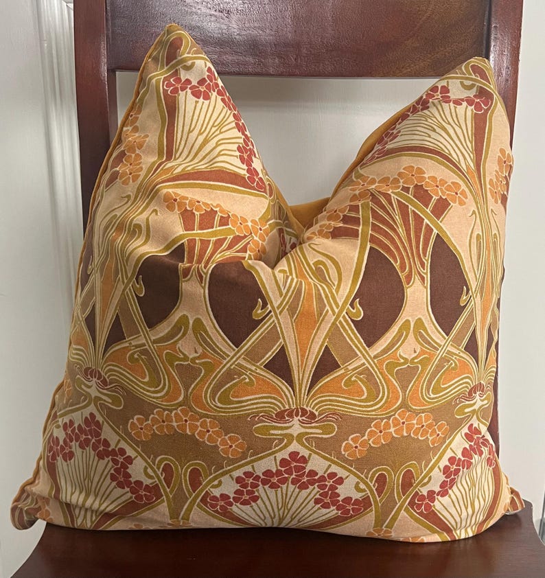 Puede incluir: Coj&iacute;n decorativo con un dise&ntilde;o floral Art Nouveau en tonos marr&oacute;n, naranja y crema. El patr&oacute;n presenta flores y hojas estilizadas, creando una est&eacute;tica vintage. El coj&iacute;n est&aacute; colocado sobre una silla de madera. Medidas: aproximadamente 40 cm x 40 cm.