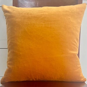 Puede incluir: Un coj&iacute;n cuadrado de terciopelo amarillo dorado. El coj&iacute;n es de color liso con un aspecto suave y texturizado. El coj&iacute;n est&aacute; sobre una superficie marr&oacute;n oscuro.