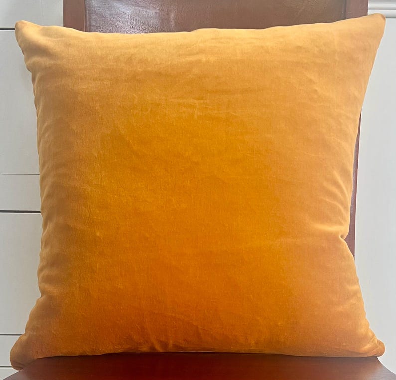 Puede incluir: Un coj&iacute;n cuadrado de terciopelo amarillo dorado. El coj&iacute;n es de color liso con un aspecto suave y texturizado. El coj&iacute;n est&aacute; sobre una superficie marr&oacute;n oscuro.