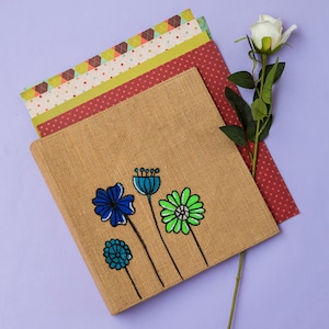 May include: A tan burlap journal cover with embroidered blue and green flowers. The journal is next to patterned paper and a white rose on a purple background. The flowers have black stems.
