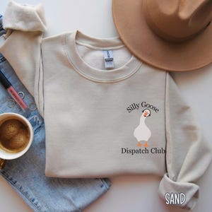 May include: A beige sweatshirt with a white goose wearing headphones and the text "Silly Goose Dispatch Club" printed on the front. The sweatshirt is laid out on a blue denim background.