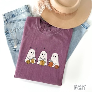 May include: A berry-colored t-shirt featuring a graphic of three cartoon ghosts knitting with yarn. The shirt is folded on top of a pair of light blue jeans, with a tan hat and decorative sprigs in the background. The word "BERRY" is printed on the bottom right.