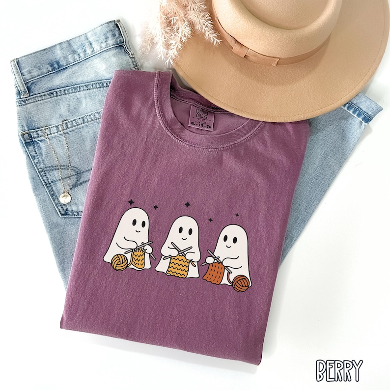 May include: A berry-colored t-shirt featuring a graphic of three cartoon ghosts knitting with yarn. The shirt is folded on top of a pair of light blue jeans, with a tan hat and decorative sprigs in the background. The word "BERRY" is printed on the bottom right.