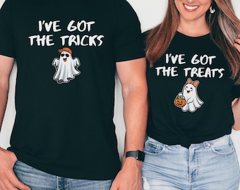May include: Two black t-shirts featuring white text and ghost graphics. The left shirt displays the text I'VE GOT THE TRICKS with a ghost wearing an orange cap and sunglasses. The right shirt displays I'VE GOT THE TREATS with a ghost wearing a bow.