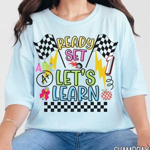 May include: Light blue t-shirt with the text "READY SET LET'S LEARN" in yellow, pink, and green. The design includes checkered flags, lightning bolts, and a race car wheel. A number 1 is also featured in the design.