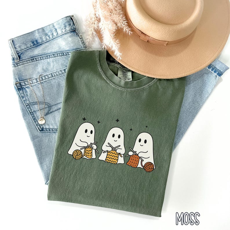 May include: A moss green t-shirt featuring three cartoon ghosts knitting with yarn. The shirt is laid on top of light wash denim jeans and a tan fedora hat. A gold necklace and dried flowers are also visible.