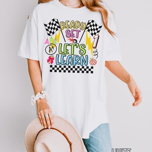 May include: White t-shirt with a colorful graphic that says "Ready Set Let's Learn". The design includes checkered flags, lightning bolts, and an apple. The shirt is a comfortable, oversized fit.