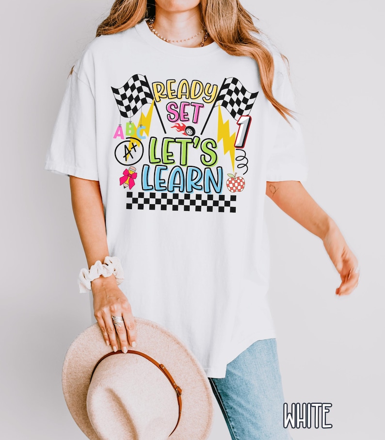 May include: White t-shirt with a colorful graphic that says "Ready Set Let's Learn". The design includes checkered flags, lightning bolts, and an apple. The shirt is a comfortable, oversized fit.