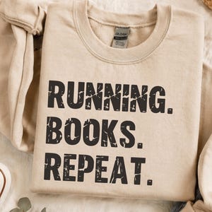 May include: A beige crewneck sweatshirt featuring the black distressed text RUNNING. BOOKS. REPEAT. printed on the front. The garment has a ribbed collar and long sleeves, with a Gildan brand label visible inside the neckline of the shirt.