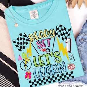 May include: A light blue t-shirt with the words "READY SET LET'S LEARN" in a colorful design. The shirt features checkered flags, lightning bolts, and an apple. The text "CHALKY MINT" is at the bottom.