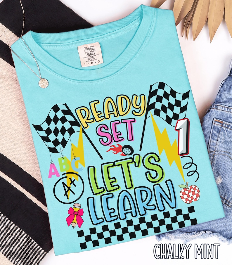 May include: A light blue t-shirt with the words "READY SET LET'S LEARN" in a colorful design. The shirt features checkered flags, lightning bolts, and an apple. The text "CHALKY MINT" is at the bottom.