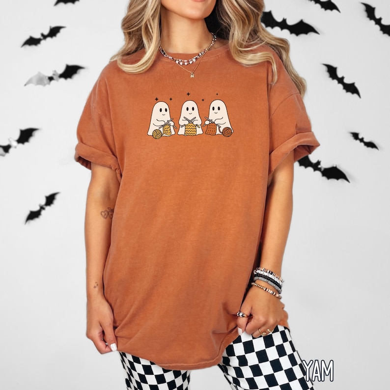 May include: A rust-colored t-shirt featuring a graphic of three friendly ghosts holding yarn baskets. The ghosts have simple, smiling faces and are adorned with small stars above them. The shirt has short sleeves and a relaxed fit, perfect for Halloween.