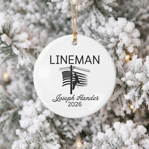 May include: A round white ceramic ornament featuring the black text LINEMAN at the top, a graphic of a utility worker on a pole with an American flag, and the personalized black text Joseph Hander 2026 printed at the bottom.