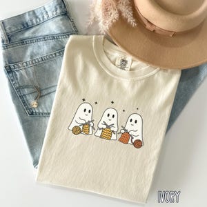 May include: Ivory-colored t-shirt featuring a graphic of three cartoon ghosts knitting with yarn. The shirt is laid on top of blue jeans, a tan hat, and a silver necklace. The text "IVORY" is printed at the bottom of the shirt.